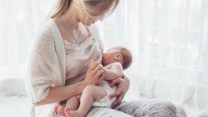 Mãe sentada junto a uma janela de cortinado claro, a amamentar o bebé ao colo
