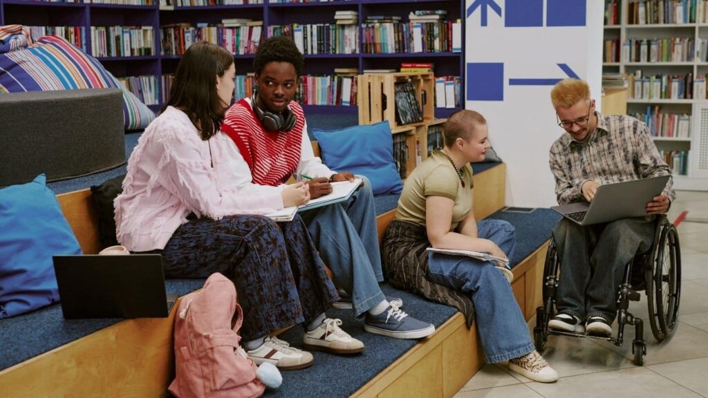 Estudantes reunidos numa biblioteca universitária, entre estantes de livros, a trabalhar em equipa — um deles em cadeira de rodas, com portátil