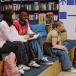 Estudantes reunidos numa biblioteca universitária, entre estantes de livros, a trabalhar em equipa — um deles em cadeira de rodas, com portátil