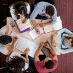 Vista de cima de seis estudantes do ensino superior a estudar em conjunto à volta de uma mesa, com livros, cadernos e tablets