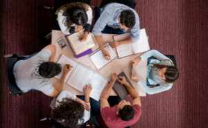 Vista de cima de seis estudantes do ensino superior a estudar em conjunto à volta de uma mesa, com livros, cadernos e tablets